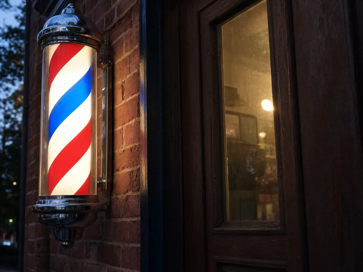 A lit red, white and blue barber pole beside a wooden shop door at dusk